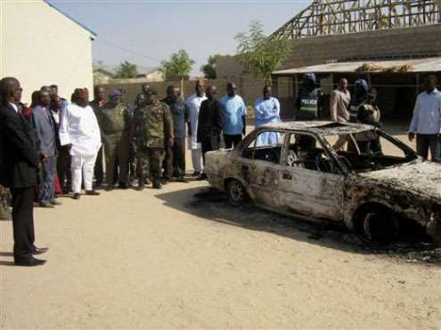 Bystanders gather around a burned car outside the Victory Baptist Church in Maiduguri, Nigeria, Saturday, Dec. 25, 2010. Authorities say dozens of assailants attacked the church on Christmas Eve, killing the pastor, two members of the choir and two people passing by the church. Police are blaming members of Boko Haram, a radical Muslim sect. (AP Photo – Njadvara Musa)