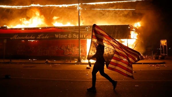 A protester in Minneapolis carries America’s flag upside down as a building burns